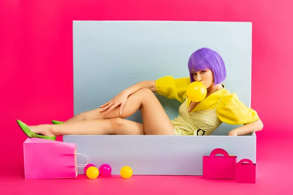 Sexy pop art girl in purple wig as doll blowing bubble gum while sitting in blue box with balls and shopping bags, on pink — Stock Photo