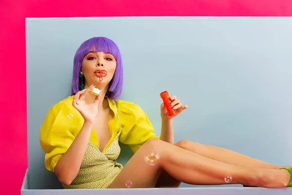 Menina bonita em peruca roxa como boneca soprando bolhas de sabão enquanto sentado na caixa azul, isolado em rosa — Stock Photo