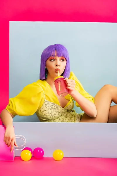 Pop art girl in purple wig as doll drinking from jar while sitting in blue box with balls and shopping bag, on pink — Stock Photo