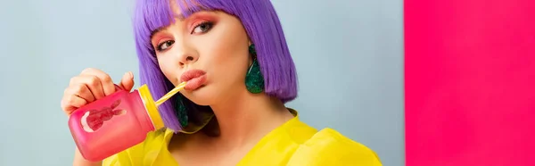 Panoramic shot of bored girl in purple wig as doll drinking from jar while sitting in blue box, isolated on pink — Stock Photo