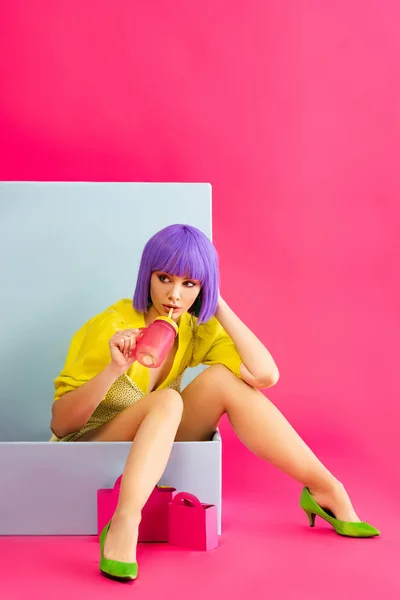 Bella ragazza pop art in parrucca viola come bambola che beve dal barattolo mentre seduto in scatola blu con borse della spesa, su rosa — Foto stock