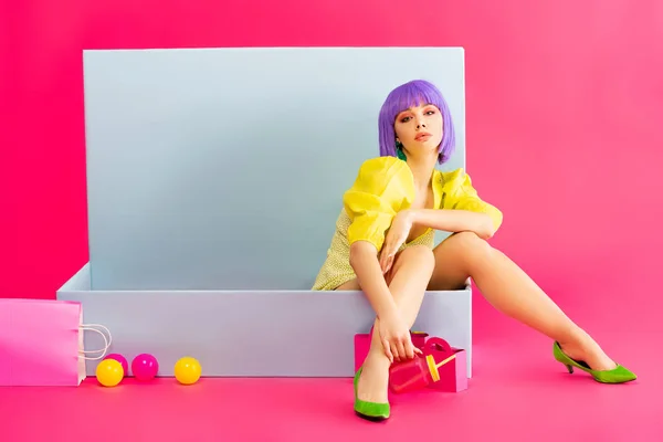 Bored girl in purple wig as doll sitting in blue box with balls and shopping bags, on pink — Stock Photo