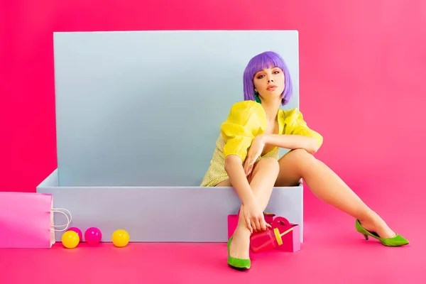 Fille ennuyée en perruque violette comme poupée assise dans une boîte bleue avec des boules et des sacs à provisions, sur rose — Photo de stock