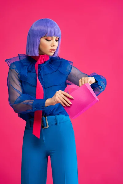 Beautiful stylish girl in purple wig holding little shopping bag, isolated on pink — Stock Photo