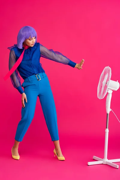 Atractiva chica con estilo en peluca púrpura de pie cerca de ventilador eléctrico, en rosa — Stock Photo