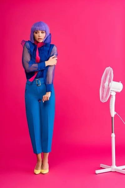 Cold stylish girl in purple wig standing near electric fan, on pink — Stock Photo