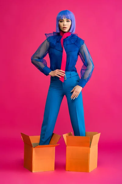Fashionable girl in purple wig standing in cardboard boxes on pink — Stock Photo