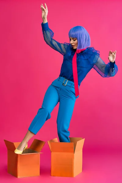 Beautiful girl in purple wig standing in cardboard boxes on pink — Stock Photo