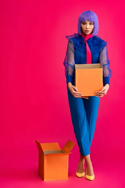 Attractive girl in trendy purple wig standing in cardboard boxes on pink — Stock Photo