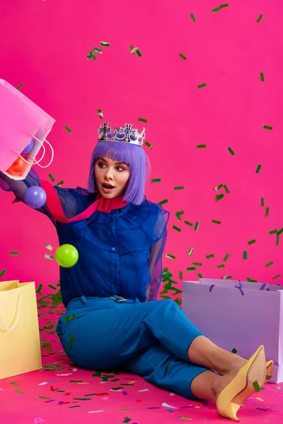 Femme choquée en perruque violette et couronne assise avec des sacs à provisions, des boules et des confettis de vacances, sur rose — Photo de stock