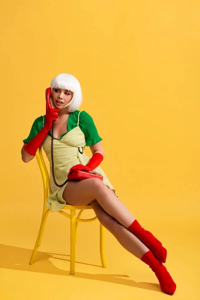 Pop art femme en perruque blanche parlant sur téléphone rétro tout en étant assis sur chaise, sur jaune — Photo de stock