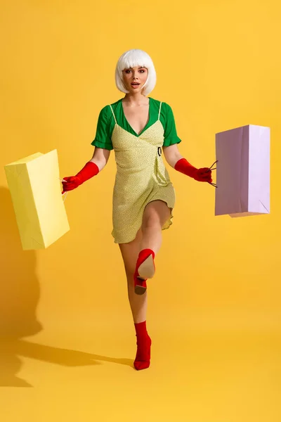 Überraschtes Pop-Art-Mädchen in weißer Perücke, das mit Einkaufstaschen spaziert, auf gelb — Stockfoto
