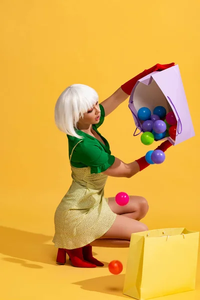 Surprised girl in white wig holding shopping bags with colorful balls, on yellow — Stock Photo