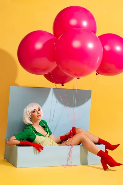 Schönes Mädchen mit weißer Perücke als Puppe in blauer Schachtel sitzend und Luftballons haltend, auf gelb — Stockfoto