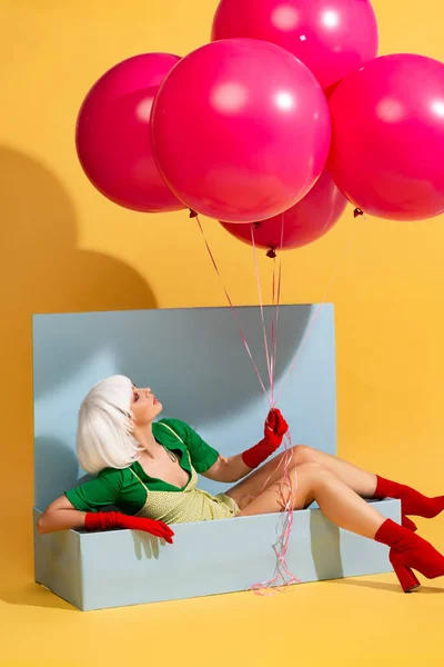 Attractive pop art girl in white wig as doll sitting in blue box and holding balloons, on yellow — Stock Photo