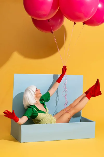 Excited pop art girl in white wig as doll sitting in blue box and holding balloons, on yellow — Stock Photo