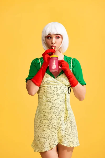Überraschtes Pop-Art-Mädchen in gelbem Kleid und weißer Perücke trinkt frisches Getränk aus Glas, isoliert auf gelb — Stockfoto
