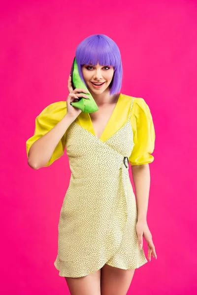Счастливая девушка поп-арта в фиолетовом парике, держащая зеленую обувь в качестве телефона, изолированная на розовом — стоковое фото