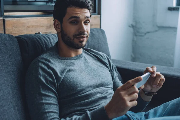 Hombre guapo mirando a la cámara mientras ve porno en el teléfono inteligente en el sofá - foto de stock