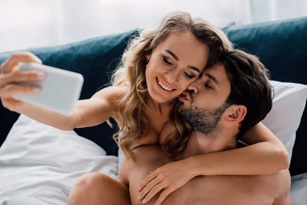 Homem muscular beijando sorridente namorada enquanto tomar selfie na cama — Fotografia de Stock