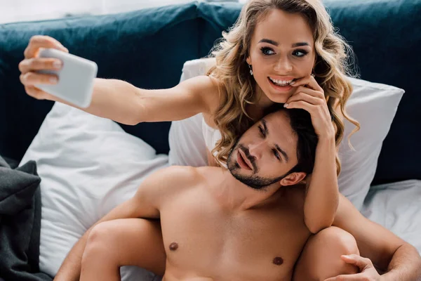 Hermosa mujer sonriente tomando selfie con teléfono inteligente cerca de novio muscular en la cama - foto de stock