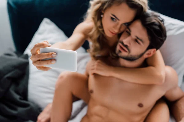 Selective focus of beautiful woman hugging muscular boyfriend while taking selfie with smartphone on bed isolated on grey — Stock Photo