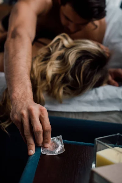 Selektiver Fokus des Mannes, der Kondom vom Nachttisch nimmt, während er mit Freundin im Bett liegt — Stockfoto
