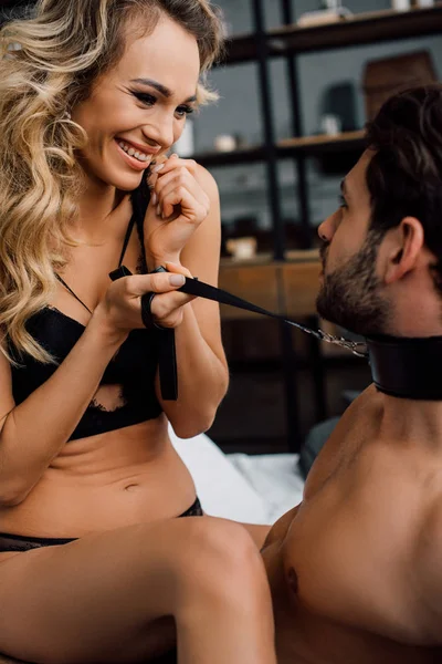 Sexy woman smiling at boyfriend while holding bdsm leash on bed — Stock Photo