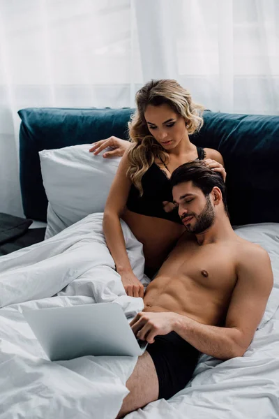 Hombre muscular mirando portátil cerca de mujer atractiva en la cama - foto de stock