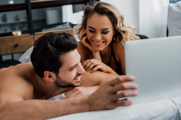 Selective focus of handsome man lying near beautiful smiling woman and laptop on bed — Stock Photo