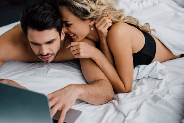 Femme souriante couchée près de bel homme utilisant un ordinateur portable sur le lit isolé sur noir — Photo de stock