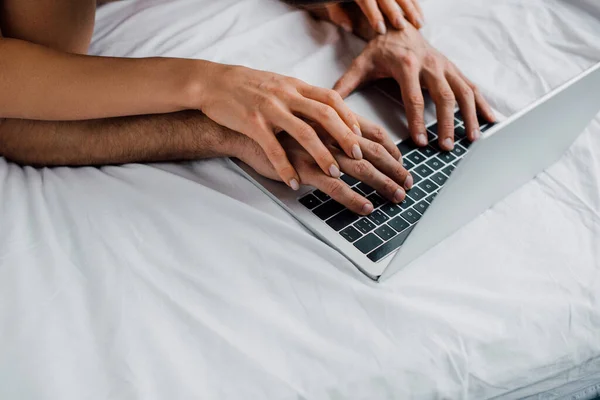 Vista recortada de la mujer tocando las manos del novio mientras usa el ordenador portátil en la cama - foto de stock