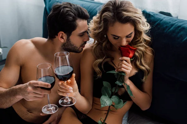 Beautiful woman smelling rose and toasting red wine with boyfriend on bed — Stock Photo