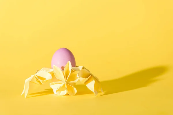 Цветы оригами рядом с окрашенным пасхальным яйцом на желтом с местом для копирования — стоковое фото
