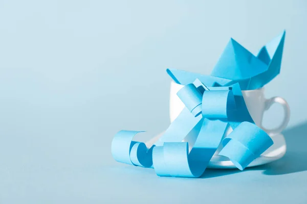 Paper ship in white cup near ribbons on blue — Stock Photo