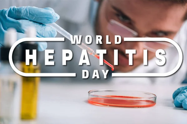 Enfoque selectivo del científico que sostiene la pipeta con líquido rojo en el laboratorio con ilustración del día mundial de la hepatitis - foto de stock