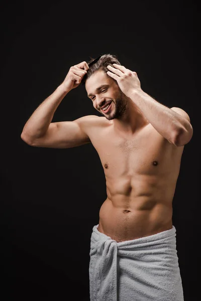 Cheerful handsome man in towel combing hair isolated on black — Stock Photo