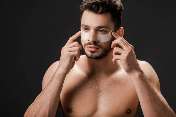 Séduisant nu homme appliquant des patchs pour les yeux sur le visage isolé sur noir — Photo de stock