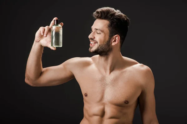 Sexy sonriente hombre desnudo sosteniendo botella de colonia aislado en negro - foto de stock