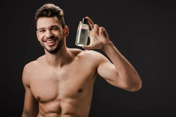 Sexy positivo desnudo hombre celebración botella de colonia aislado en negro - foto de stock