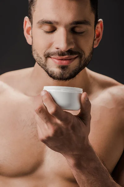 Positive naked man holding plastic container with face cream isolated on black — Stock Photo
