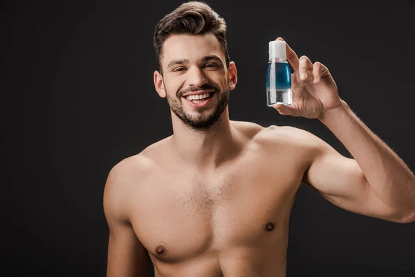 Sonriente sexy desnudo hombre celebración botella con cosmético líquido aislado en negro - foto de stock