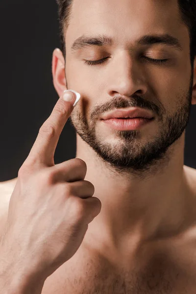 Homem nu com olhos fechados aplicando creme facial isolado em preto — Fotografia de Stock