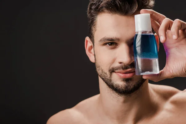 Bonito sorridente homem segurando garrafa com líquido cosmético isolado no preto — Fotografia de Stock