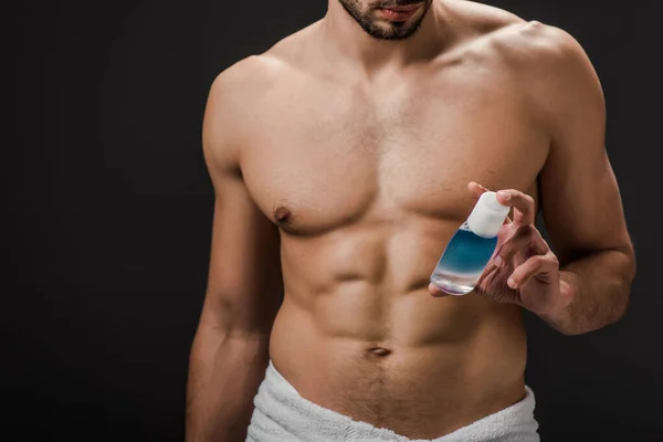 Обрезанный вид сексуального мужчины без рубашки, держащего бутылку с косметической жидкостью, изолированной на черном — стоковое фото