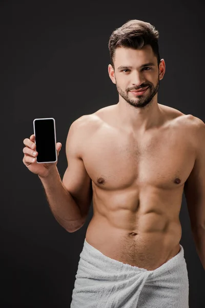 Улыбающийся сексуальный мужчина в полотенце, показывающий смартфон с пустым экраном, изолированным на черном — стоковое фото