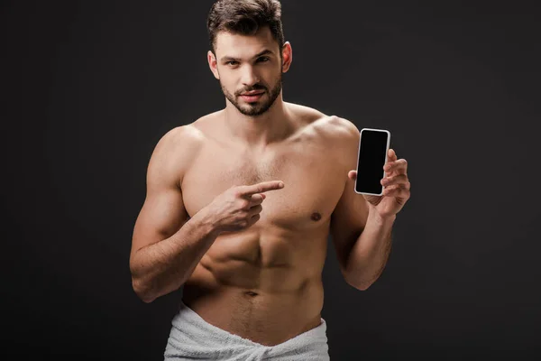 Sexy man in towel pointing at smartphone with blank screen isolated on grey — Stock Photo