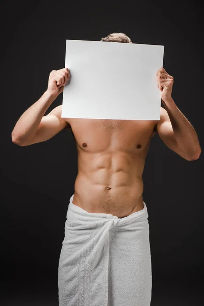 Sexy sin camisa hombre en toalla sosteniendo en blanco cartel en frente de la cara aislado en negro - foto de stock