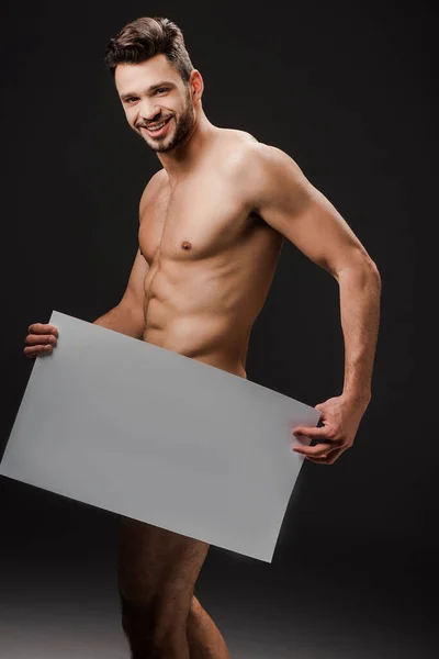 Smiling sexy naked man holding blank placard isolated on black — Stock Photo