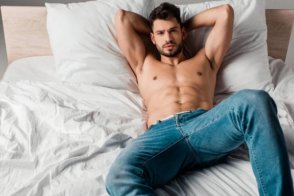 Sexy shirtless macho in jeans lying on bed on grey — Stock Photo